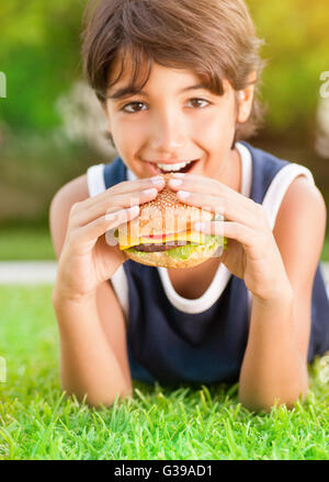 Portrait of a cute happy teen boy lying down on fresh green grass and with pleasure eating burger, tasty, but unhealthy food Stock Photo