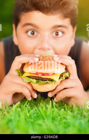 Portrait of a hungry teen boy lying down on fresh green grass and with pleasure eating burger, enjoying picnic outdoors Stock Photo