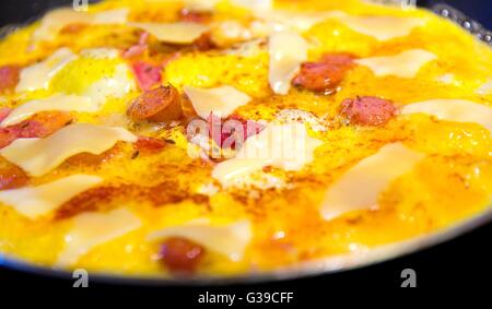 Frying ham and eggs with cheese, sausages and pepper in pan sprinkled by caraway seeds closeup. Stock Photo