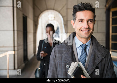 Smiling corporate businessman carrying newspaper and cell phone in cloister Stock Photo
