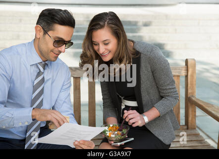Corporate businessman and businesswoman eating lunch and reviewing paperwork on bench Stock Photo