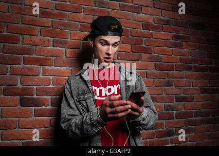 Young surprised guy looking at smart phone and listening to music against brick wall Stock Photo