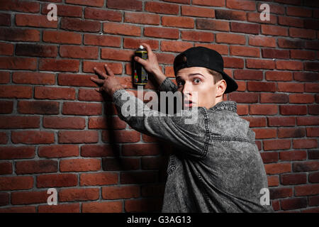 Graffiti man in shirt spraying red brick wall by aerosol can Stock Photo