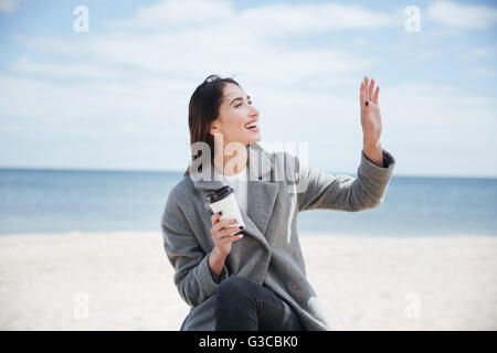 Cheerful pretty attractive woman waving to someoone and holding take away cup at the seaside Stock Photo
