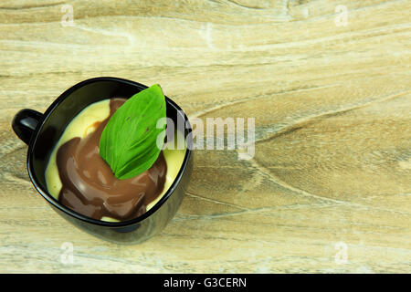 Chocolate-vanilla pudding in a black cup decorated with fresh basil leaf on wooden background. Horizontal , flat,top view. Stock Photo