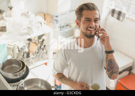 Tattooed young man talking on cell phone and drinking coffee in apartment kitchen Stock Photo