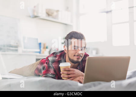 Young man drinking coffee at laptop on bed Stock Photo