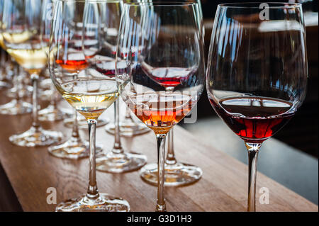 Three wine glasses with white, rose, and red wine samples, on wood counter with other glasses in background.