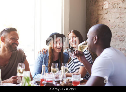 Two attractive ladies in hat and striped shirt laughing while embarrassed male friends drink wine in restaurant with large brigh Stock Photo