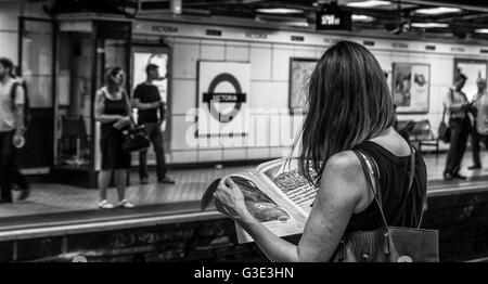 Woman reading a newspaper on the platform of Victoria Underground Station, whilst waiting for a train, London, UK Stock Photo