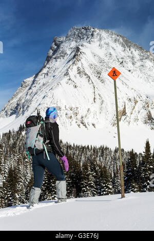 Female snowshoer on snow covered hillside with snow covered mountains and trees in the background with snowshoe sign and blue sky with clouds Stock Photo