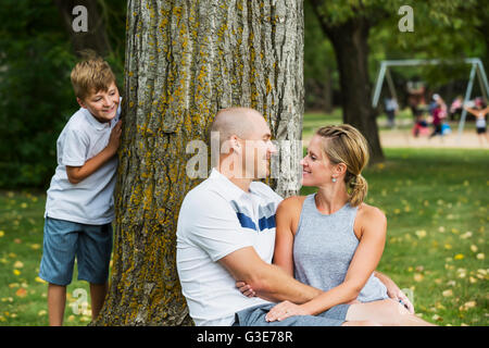 A married couple spending quality time together in a park during a family outing with their son peeking from behind a tree; Edmonton, Alberta, Canada Stock Photo