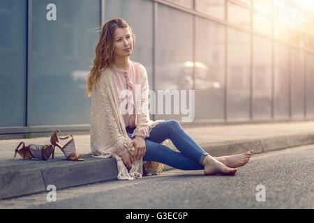 Serious pretty trendy young woman sitting relaxing on a sidewalk on an urban street in her high heels and fashionable outfit Stock Photo