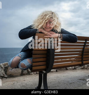 Beautiful blonde girl with curly hair in ripped jeans is seating on the bench near of the sea Stock Photo