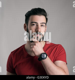 Puzzled young man stroking touching beard looking at camera over gray background Stock Photo