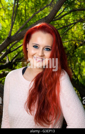 A beautiful, teenage girl with dyed red hair poses for a portrait in front of trees.  She has a tilted head, lovely smile, and Stock Photo