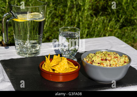 Home made guacamole in ceramic bowl and tortilla chips and soured cream on the side with jug and glass of iced water. On slate m