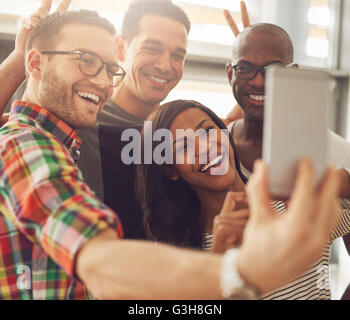 Group of four diverse cheerful co-workers taking self portrait and making funny gestures with hands at small office in front of Stock Photo