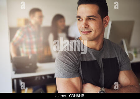 Close up of calm handsome male worker with folded muscular arms wearing gray short sleeve shirt in small office with other emplo Stock Photo
