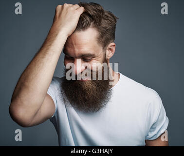 Laughing young man with long beard and white shirt holding hair while facing downward over gray background Stock Photo