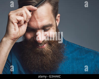 Well groomed handsome bearded man in blue denim shirt and happy expression combing his hair over gray background Stock Photo