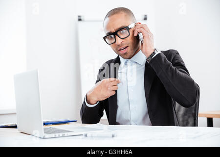 Portrait of a surprised businessman talking on mobile phone at working place in office Stock Photo