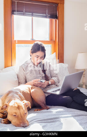 Woman sitting on bed with dog and laptop using smartphone Stock Photo