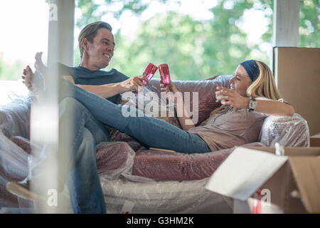 Moving house: couple relaxing on bubbled wrapped sofa, in room with cardboard boxes, holding champagne flutes, making a toast Stock Photo