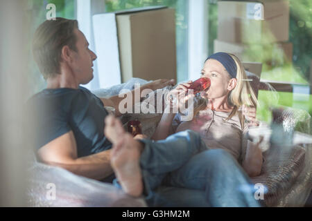 Moving house: couple relaxing on bubbled wrapped sofa, in room with cardboard boxes, drinking from champagne flutes Stock Photo