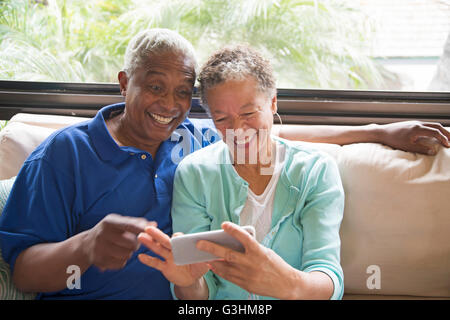 Senior couple sitting on sofa, looking at smartphone, laughing Stock Photo