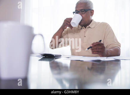 Senior man sitting at table, drinking hot drink, doing paperwork Stock Photo
