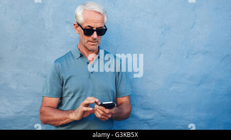 Portrait of mature man in sunglasses reading text message on her mobile phone against blue background. Middle aged caucasian mal Stock Photo