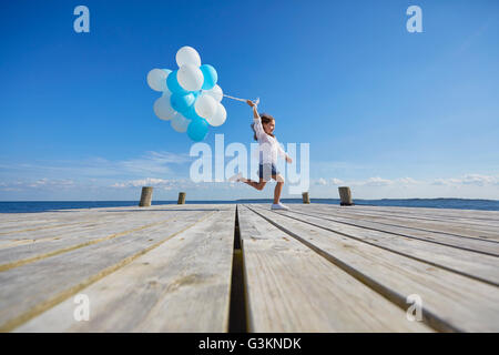 Young girl running on wooden pier, holding bunch of balloons Stock Photo
