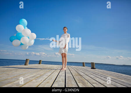 Portrait of young woman, standing on wooden pier, holding bunch of balloons Stock Photo
