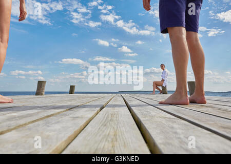 Young woman sitting on post on wooden pier, looking at friends standing further away Stock Photo