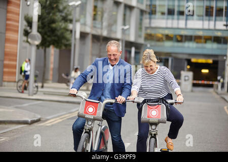 Mature dating couple laughing whilst cycling on hire bicycles, London, UK Stock Photo