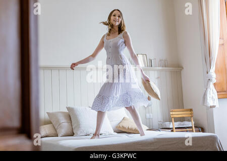 Young woman in long white dress jumping on bed looking at camera smiling Stock Photo