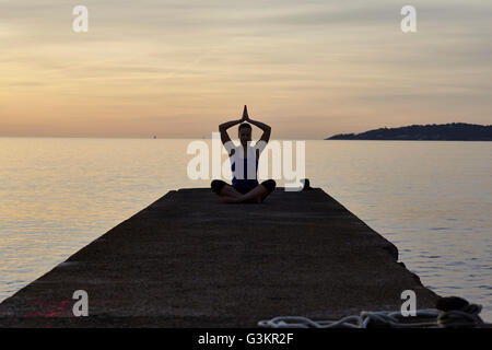 Young woman sitting on pier, in yoga position, at dusk Stock Photo