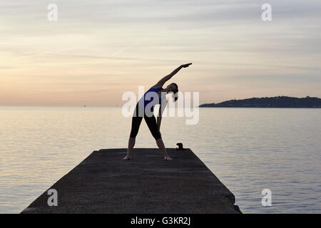 Young woman standing on pier, in yoga position, at sunset Stock Photo