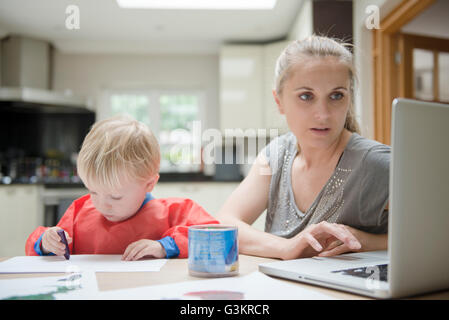Mother using laptop while son draws on paper beside her Stock Photo