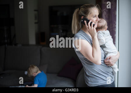 Mother holding baby boy, speaking on smartphone, while son uses digital tablet Stock Photo