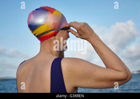 Mature woman wearing swimming costume and swimming hat, standing beside the sea, adjusting swimming goggles, rear view Stock Photo