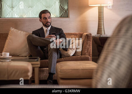 Young businessman sitting on hotel lobby sofa using smartphone Stock Photo