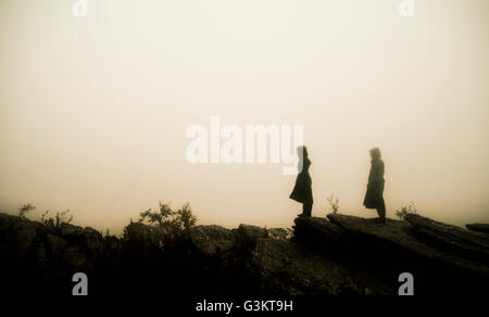 Side view of couple wearing raincoats standing on rocks in fog looking away Stock Photo