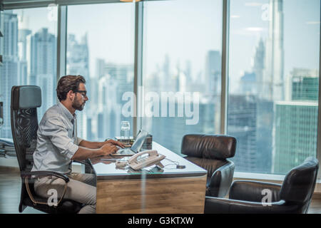 Businessman using laptop at desk with window view of Burj Khalifa, Dubai, United Arab Emirates Stock Photo