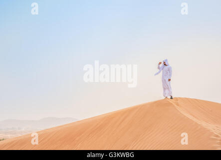 Middle eastern man wearing traditional clothes looking out from desert dune, Dubai, United Arab Emirates Stock Photo