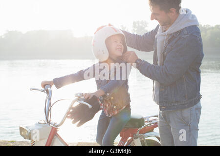 Young girl sitting on father's moped, father putting crash helmet on daughter's head, smiling Stock Photo