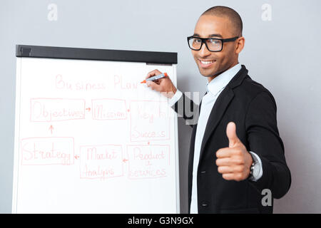 Happy african young man in glasses writing on flipchart and showing thumbs up Stock Photo