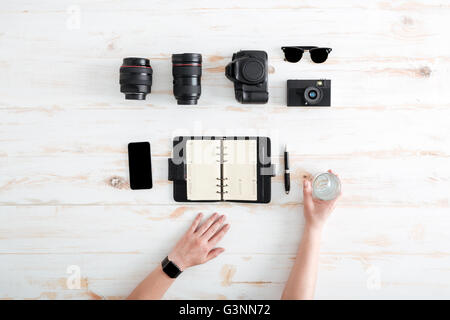 Hands of young man writing in notebook and drinking water on wooden table with blank screen mobile phone, sunglasses, lenses and Stock Photo