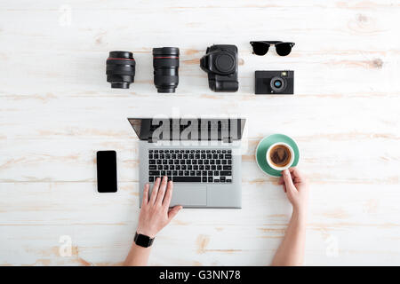 Hands of young man using laptop and drinking coffee on wooden table with blank screen mobile phone, sunglasses, lenses and photo Stock Photo